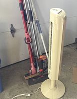 Side view of cleaning mops with varied handles and heads standing next to vertical tower fan in a garage setting.