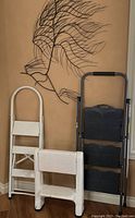 Three step ladders of various types and a metal wall hanging mounted on wall behind them.