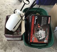 Photo of two items: a boxed Craftsman 19.2 Volt cordless 5.5 inch trim saw and a white handheld sprayer with a black wand, placed inside storage bins on a concrete floor