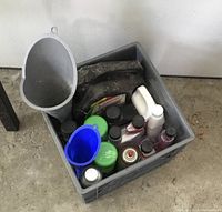 Top view of plastic bin containing multiple used automotive fluid bottles and three plastic funnels in gray, blue, and black.