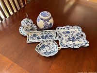 Photo of five blue and white Vienna porcelain hostess serving pieces on a wooden table: jam jar with lid and spoon, oblong chocolate tray, scalloped cookie plate, sauce pitcher, and ginger jar with lid.
