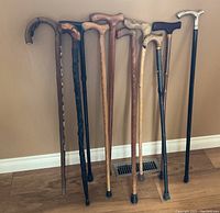 Photo showing eight assorted walking canes standing against a wall on a hardwood floor, featuring carved wooden designs and metal adjustable canes with varied handle styles and rubber tips.