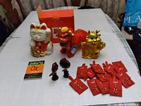 Photo showing three Asian Feng Shui statues including a white Maneki-neko, a red lion dance figure, and a gold cat, along with red cloth pouches, small dark amulets, and a Feng Shui booklet.