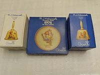 Photo showing original boxes of two Hummel bells and one collector plate arranged side by side on a tabletop.