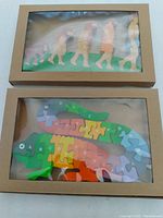 Top view of two wooden puzzles in cardboard boxes with plastic windows. Human evolution puzzle on top shows 5 evolution stages in pastel colors. Iguana puzzle with alphabet-labeled pieces in multiple colors below.
