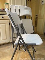 Set of four metal folding chairs stacked together, showing white plastic seats and backs with grey metal frames