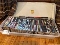 Wide view of the plastic bin filled with numerous jewel CD cases arranged standing upright inside with a white lid behind it.