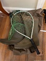 Photo of olive green chest waders with black boots and fishing net with green mesh and silver frame on wooden floor.