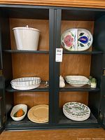 Closed cabinet shelf with white ceramic soup tureen with lid (top left), oval divided floral tray (top right), beige and green botanical plates, pie dishes, and salt and pepper shakers arranged on shelves.