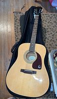 Full view of the Epiphone acoustic guitar with light wood finish and black soft case underneath.