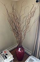 Full view of tall faux plant decor in a corner, showing thin decorative wood branches extending upward from a red ceramic planter.