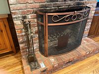 Photo showing copper-colored fireplace screen with decorative scroll design and black metal mesh screen, with fireplace tool holder and tools beside.