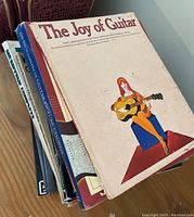 Books stacked on top of each other, with visible titles The Joy of Guitar, The Encyclopedia of Guitar Picture Chords, and others.