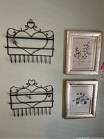Photo shows two black metal jewelry hangers with heart design and multiple hooks, alongside two small silver framed prints with botanical illustrations.