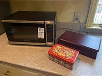Black and Decker microwave oven and two storage boxes on countertop. Boxes are Boston Tea Company wooden box and La Cure Gourmande decorative metal box.