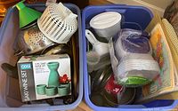 Two large blue plastic bins filled with various kitchen items including pots, pans, colanders, plastic containers, cutting boards, and placemats.