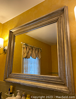 Full view of the large rectangular mirror with a thick distressed silver wooden frame hanging on a yellow wall above a sink area.