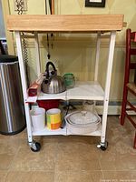 Front view of rolling kitchen table showing wooden top, two lower white shelves, caster wheels, and some kitchen items on shelves.