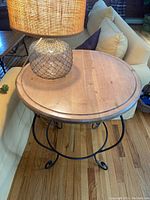 Round wooden top small table with a black metal base, placed on wooden floor next to a sofa with decorative lamp on top.
