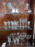 Glass stemware on two glass shelves inside wooden cabinet. Top shelf has cocktail and wine glasses, middle shelf shows smaller cordial glasses and wine glasses, bottom shelf partially visible with variety of coupe and stemmed glasses.