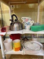 Kitchen shelf holding stainless steel kettle, floral teacups, plates, Tupperware, mugs, and bowls