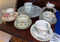 Six assorted teacups with matching saucers and three bread plates arranged on a wooden surface showing decorative floral and gold details.