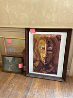 Three framed pieces of wall art leaning against wall on wooden floor, including relief portrait, landscape print, and small painting.