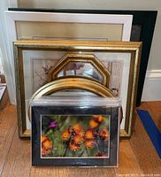 Several framed items including nature photograph, floral paintings, and mirrors stacked against a wall on floor.