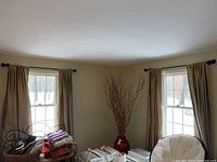 Living room setting showing two windows with beige lined curtains and dark curtain rods mounted above. Curtains hang to floor length. Piles of fabric and miscellaneous items on furniture below windows.