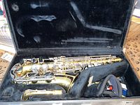 Full view of alto saxophone body and bell in black plush-lined hard case, neck strap and small accessories visible; mouthpiece absent