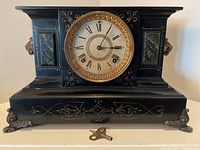 Front view of black mantel clock showing circular Roman numeral dial, gold-tone bezel, decorative painted details, sitting on scrolled metal feet with winding key in front