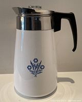 Front view of white Corning Ware coffee pot with black handle, stainless steel lid, and blue floral design at the front.