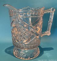 Front and side views of a clear pressed glass pitcher with starburst and daisy motifs, scalloped top rim, and decorative handle.
