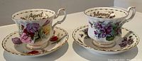 Side view of February and April floral tea cups and saucers showing detailed flower patterns and gold trim.