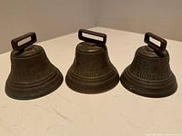 Three cast metal antique bells with rectangular handles arranged in a row showing front detail and inscriptions.