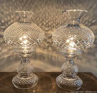 Two clear Waterford crystal lamps with diamond cut crystal pattern, lit and casting light patterns on the wall. Both lamps composed of two pieces each.