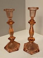 Pair of pink Depression glass candle holders shown side by side on a white surface against a neutral background, highlighting the front and side views of the faceted, ribbed design statuettes with hexagonal bases.