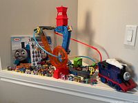 Full view of assembled Thomas the Train motorized playset including track, tower, and orange rotating component, with 39 mini Thomas train cars arranged in front, remote control Thomas car, encyclopedia, and sticker book in background.