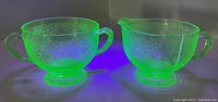 Two-piece uranium glass cream and sugar set glowing bright green under UV light, displaying etched floral patterns.