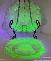 Pair of uranium glass octagonal dinner plates displayed on black stands showing detailed sunburst and floral patterns illuminated by UV light.