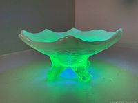 Photo under UV light showing strong green fluorescence of Vaseline glass dish, highlighting footed base and ruffled edge.