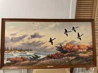 Frontal view showing the framed painting of geese flying near a coastal shoreline with autumn trees and cloudy sky.