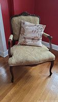 Full front view of the carved wood trim accent chair with tan embroidered leaf print fabric and floral pillow, showing the overall shape, cushion, and pillow.