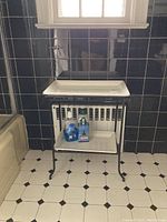 Front view of the two tier bathroom rack on black tile floor against black tiled wall, showing porcelain shelves and metal frame.