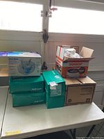 Photo showing multiple boxes on a table including green Coloplast Self-Cath boxes, a Vicks humidifier box, and other boxes likely containing bandages and wraps.