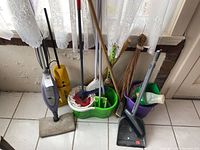 Cleaning tools including small vacuum, two mop bucket sets, brooms, a dustpan, and several brushes arranged standing near a window with lace curtains and tiled floor.