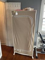Closed view of the portable clothing rack with beige fabric cover, white trim, and wheels at base, standing upright.