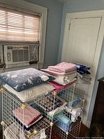 Stack of assorted linens and towels with decorative pillows on top arranged on a metal rack below a window with air conditioning unit.