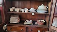Wooden cabinet with multiple shelves containing stacks of white and patterned dishware including cups, saucers, plates, bowls, and a covered serving bowl.