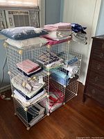 Photo of a white metal wire storage rack with cubby shelves. Various folded fabrics and pillows are displayed on the rack but are excluded from the sale.